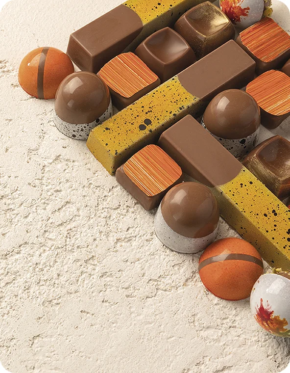 Assorted gourmet chocolates and truffles in brown, orange, and speckled yellow, arranged diagonally on a textured light surface.