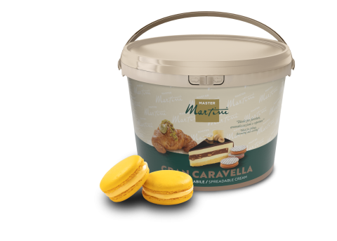 Găleată bej cu crema tartinabilă Master Martini Gran Caravella alături de două macaroane galbene și o prăjitură umplută.